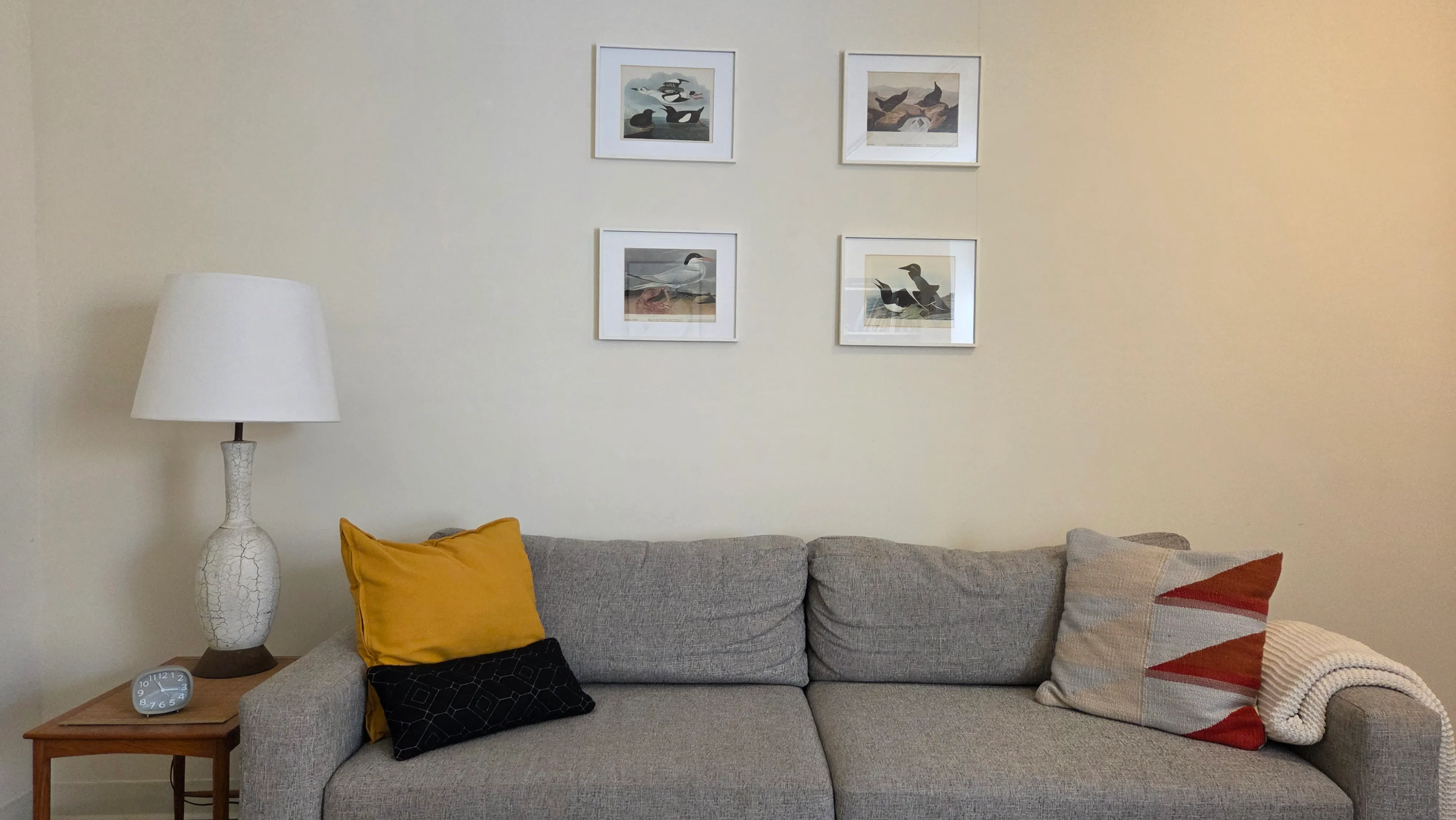 The therapy office: a grey sofa with cushions and a throw, beside a side table with a lamp and a clock, under four framed bird prints
